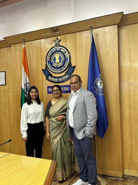 Three people standing beside the Customs emblem and the Indian flag in a wood-panelled office