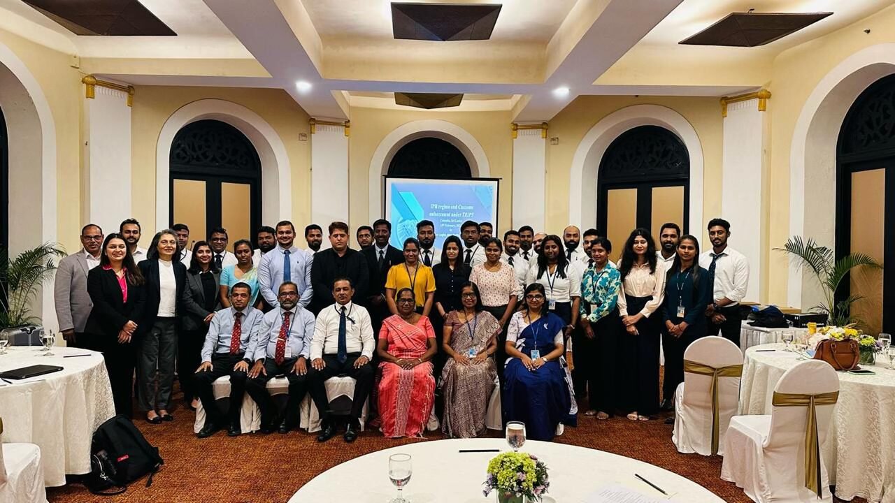 A large group of workshop participants and resource persons posing together in a hall with a presentation screen behind them